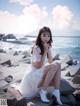 A woman in a white dress sitting on some rocks by the ocean.